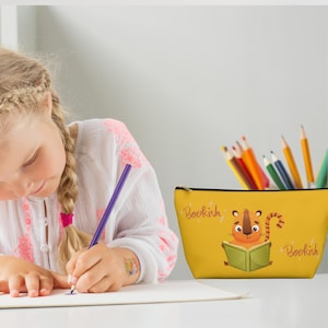May include: Yellow pencil case with a cartoon tiger reading a book. The case has the word "Bookish" printed on both sides in pink lettering.