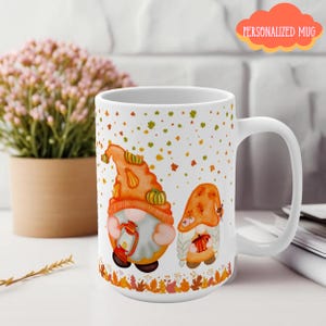 May include: White ceramic mug featuring two autumn gnomes with orange hats and pumpkins. The mug has a white handle and is decorated with falling leaves and the text "PERSONALIZED MUG". The base of the mug has a border of fall leaves.