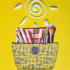 May include: A yellow and gray makeup bag with the initials "SW" in a circle. The bag is filled with makeup brushes, lip gloss, and an eyeshadow palette.