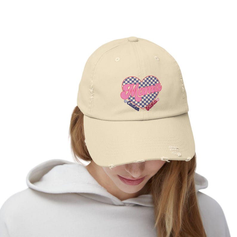 May include: A light beige distressed baseball cap with a checkered heart design and the text "Mama" in pink. The heart also includes the text "Camden" and "River" in smaller font.