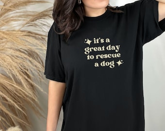 May include: Black t-shirt with the text "it's a great day to rescue a dog" in a light beige color. The text is accompanied by star graphics. The shirt is a crew neck style.