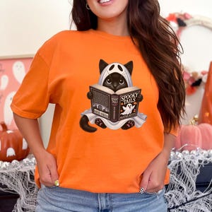 May include: Orange t-shirt with a black cat wearing a ghost costume and reading a book titled "Spooky Tails" by Agatha Kitty.