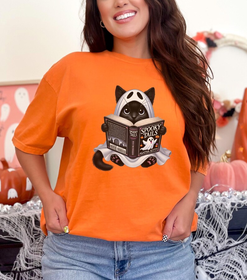 May include: Orange t-shirt with a black cat wearing a ghost costume and reading a book titled "Spooky Tails" by Agatha Kitty.