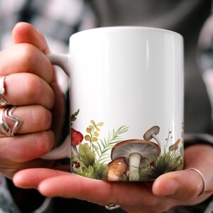 May include: White ceramic mug featuring a design of mushrooms and foliage around the base. The mug is being held by a person's hands.