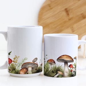 May include: Two white ceramic mugs with mushroom illustrations. The mug on the left has a white background and the mug on the right has a green and brown background with mushrooms.