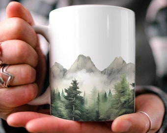 May include: White ceramic mug with a watercolor design of a misty mountain range and forest. The mug is being held by a person's hands.