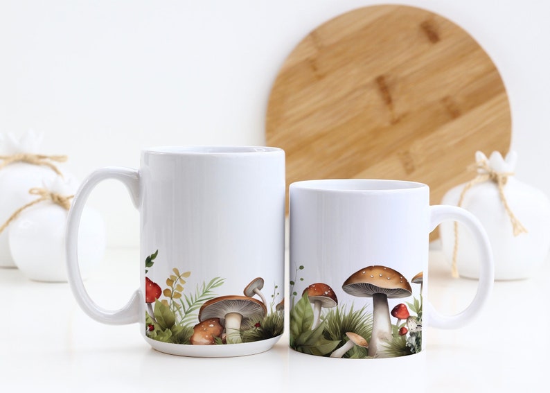 May include: Two white ceramic mugs with mushroom illustrations. The mug on the left has a white background and the mug on the right has a green and brown background with mushrooms.