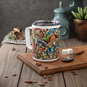 K&ouml;nnte beinhalten: Eine wei&szlig;e Keramiktasse mit einem bunten Pilzdesign. Die Tasse hat einen schwarzen Griff und einen Edelstahldeckel.