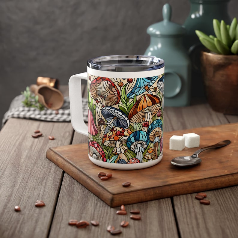 K&ouml;nnte beinhalten: Eine wei&szlig;e Keramiktasse mit einem bunten Pilzdesign. Die Tasse hat einen schwarzen Griff und einen Edelstahldeckel.