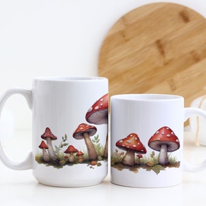 May include: Two white ceramic mugs with a design of red and white mushrooms on a white background. The mugs are on a white surface.