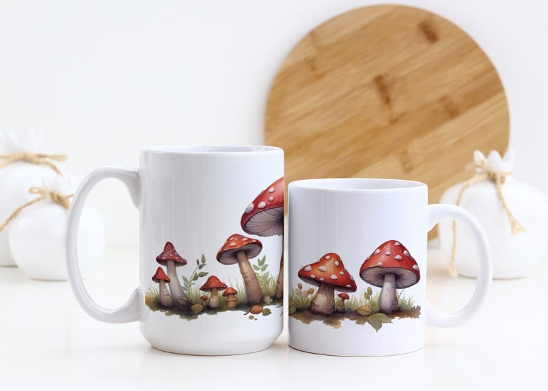 May include: Two white ceramic mugs with a design of red and white mushrooms on a white background. The mugs are on a white surface.