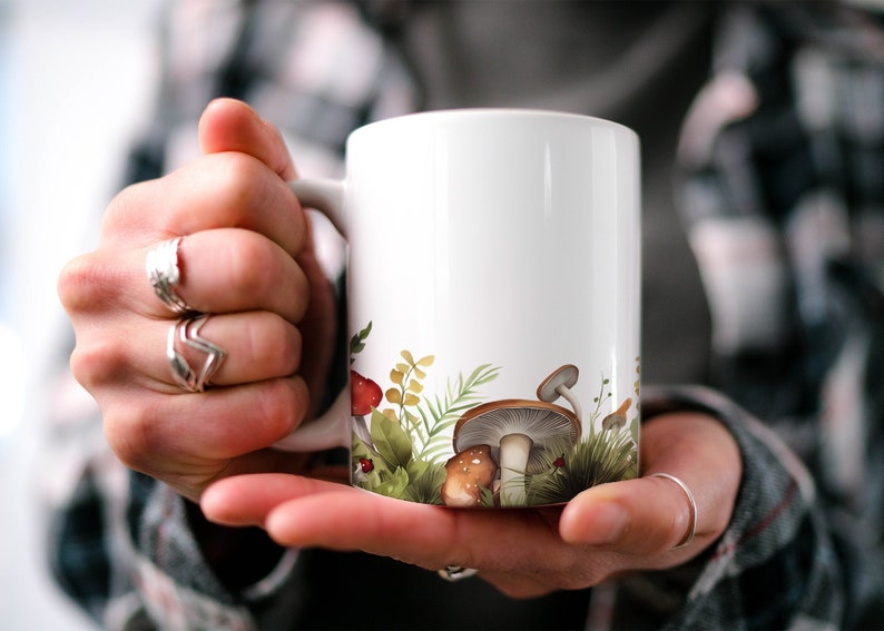 May include: White ceramic mug featuring a design of mushrooms and foliage around the base. The mug is being held by a person's hands.