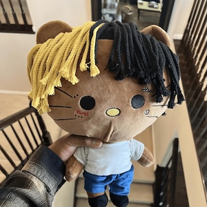 May include: A plush doll with a brown head and yellow and black yarn hair. The doll has a white shirt, blue shorts, and black shoes. The doll is being held in a person's hand.