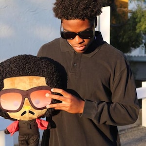 May include: A person wearing a black long-sleeved polo shirt and sunglasses is holding a plush toy. The toy is wearing sunglasses and a red shirt.