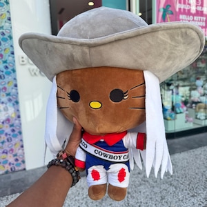 May include: A plush toy featuring a cartoon cat character wearing a gray cowboy hat and a blue, white, and red outfit. The outfit has the text "COWBOY CAT" on it. The toy has white hair and brown fur.