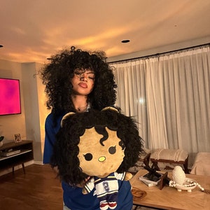 May include: A person with curly black hair is holding a plush toy. The toy is a brown and black character with a blue and yellow jersey. The jersey has the number "8" on it. The person is wearing a blue shirt and light blue jeans.