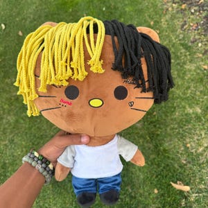 May include: A brown plush toy with yellow and black hair, wearing a white shirt and blue jeans. The toy has a Hello Kitty-like face with the words "Numb" and "Hiatus" written on it.