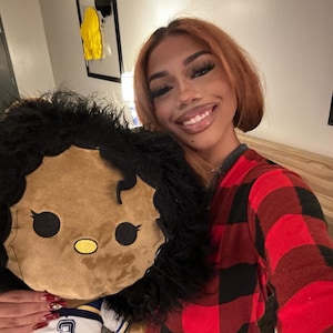 May include: A woman with long brown hair is holding a brown plush toy with black hair and a blue and yellow jersey. She is wearing a red and black plaid shirt.