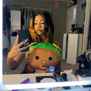 May include: A young woman with long brown hair with blonde highlights is taking a selfie in a mirror. She is holding a brown and green plush toy with a cartoon face. The toy has a power button on the front. The mirror is lit with a white light.