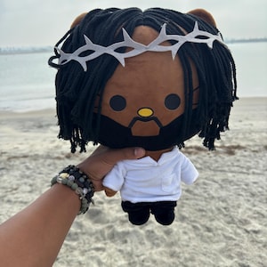Puede incluir: Una muñeca de peluche con una cara marrón, cabello negro y una corona de espinas blanca. La muñeca lleva una camisa blanca y pantalones negros. La muñeca está siendo sostenida por una persona en una playa arenosa.