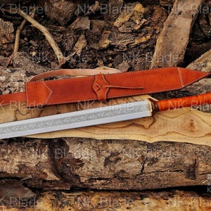 May include: A double-edged sword with a silver-toned blade featuring an intricate floral engraving along the center. The handle is wrapped in brown leather with a rounded pommel, accompanied by a matching brown leather sheath with white stitching.