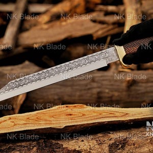 May include: A fixed-blade knife featuring a long, pointed steel blade with an intricate floral scrollwork engraving along the spine. The handle has a brown, textured, cross-hatched pattern with a polished gold-tone bolster and pommel cap.