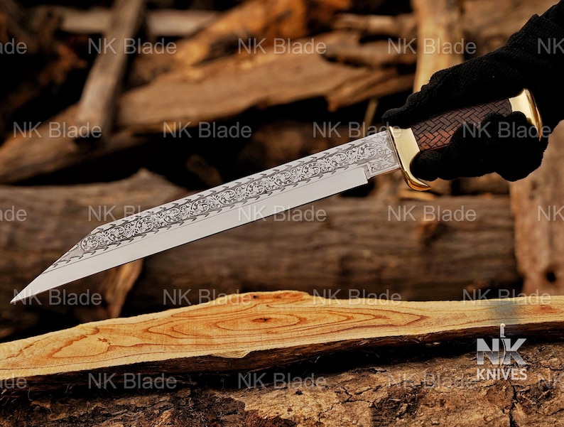 May include: A fixed-blade knife featuring a long, pointed steel blade with an intricate floral scrollwork engraving along the spine. The handle has a brown, textured, cross-hatched pattern with a polished gold-tone bolster and pommel cap.
