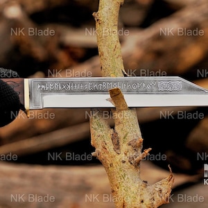 May include: A fixed-blade knife with a dark wooden handle and a metallic blade featuring etched runic inscriptions and a decorative knotwork pattern near the tip. The blade is positioned horizontally, cutting into a light brown tree branch.