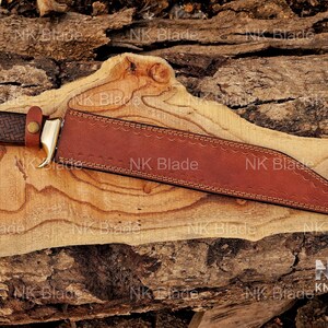 May include: A fixed-blade knife with a dark brown textured handle, a brass bolster, and a brown leather sheath. The sheath features decorative stitching along the edges and a small leather strap with a brass snap button securing the handle in place.
