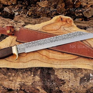 May include: A fixed-blade knife with a silver-toned steel blade featuring an intricate floral engraving along the spine. The handle is dark brown wood with a cross-hatched texture and brass-colored bolster. It includes a brown leather sheath.