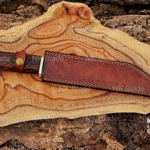 May include: A fixed-blade knife with a dark brown wooden handle featuring intricate carvings and a brass-colored snap button strap. The knife is encased in a brown leather sheath with decorative stitching along the edges and a textured surface.
