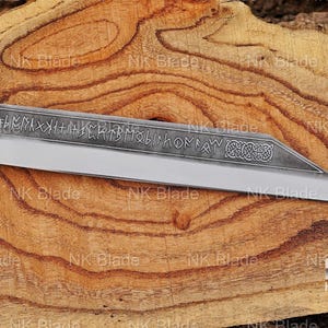 May include: A fixed-blade knife with a dark wooden handle and a brass bolster. The steel blade features an etched pattern of runic characters along the spine and a decorative Celtic knot design near the pointed tip, with a polished cutting edge.