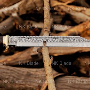 May include: A fixed-blade knife with a silver-toned blade featuring an intricate floral engraving along the spine. The handle has a dark, textured grip and a gold-toned bolster. The blade is positioned horizontally against a thin wooden branch.