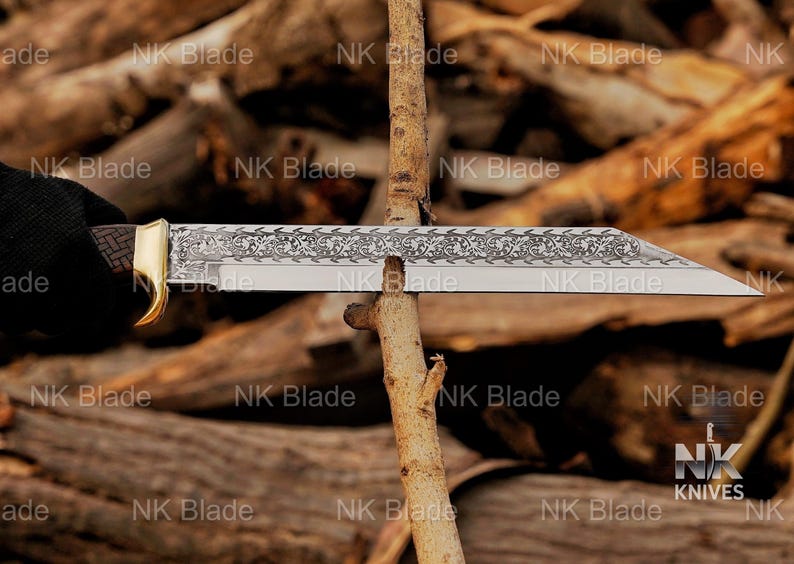 May include: A fixed-blade knife with a silver-toned blade featuring an intricate floral engraving along the spine. The handle has a dark, textured grip and a gold-toned bolster. The blade is positioned horizontally against a thin wooden branch.