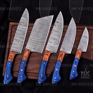 May include: A set of six kitchen knives with patterned steel blades and wooden handles. The handles are a combination of brown and blue, with brass rivets. The knives are arranged on a wooden cutting board.