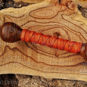 May include: A sword handle featuring a dark brown wooden pommel with gold wire inlay, a bright orange leather-wrapped grip, and a gold-toned metal bolster. The pommel is rounded, and the grip is secured with a crisscross leather cord pattern.