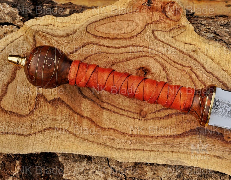 May include: A sword handle featuring a dark brown wooden pommel with gold wire inlay, a bright orange leather-wrapped grip, and a gold-toned metal bolster. The pommel is rounded, and the grip is secured with a crisscross leather cord pattern.
