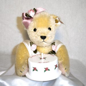 May include: A plush teddy bear with a pink bow and roses, holding a white cake. The cake is decorated with red roses and the text "Happy Takes the Cake 30 Years". The bear is wearing a white dress with rose accents.