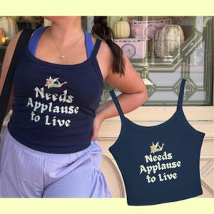 May include: A navy blue tank top featuring a graphic of a small fairy character and the text "Needs Applause to Live" printed in a light cream color. The garment has thin shoulder straps and a scoop neckline, presented as a sleeveless top.