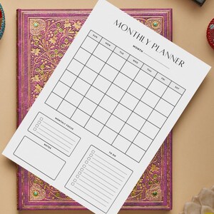 May include: A white monthly planner sheet with a grid layout, labeled "MONTHLY PLANNER." The sheet includes sections for the month, days of the week, monthly focus, notes, and to-do lists. It rests on a purple journal with gold floral designs.