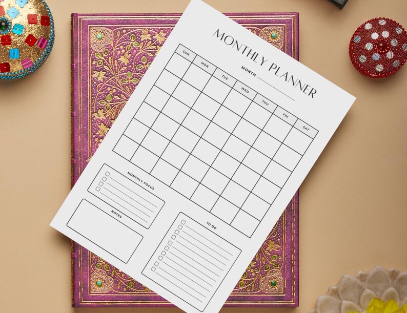 May include: A white monthly planner sheet with a grid layout, labeled "MONTHLY PLANNER." The sheet includes sections for the month, days of the week, monthly focus, notes, and to-do lists. It rests on a purple journal with gold floral designs.