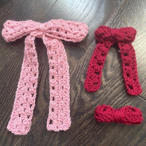 May include: Two crochet bow ties in shades of pink and red. The larger pink bow tie has long, dangling ends. The smaller bow ties are a deep red. The items are handmade and appear to be made of yarn.