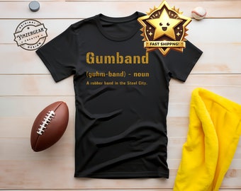 Op de afbeelding: Zwart T-shirt met gouden tekst die "Gumband (guhm-band) - zelfstandig naamwoord Een elastiekje in de stalen stad." luidt.