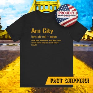 May include: Black t-shirt with the text "Arn City" in gold lettering. The text includes a definition of the phrase and a description of local beer. A round sticker with the American flag is on the upper right.