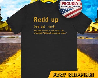 May include: Black t-shirt with gold text that reads "Redd up (red-up) verb Any kind of soda or soft drink. The preferred Pittsburgh term over "soda.""