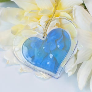 May include: A blue glass heart ornament with a white flower background. The heart is clear and has a blue design inside.