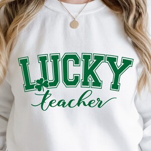 K&ouml;nnte beinhalten: Wei&szlig;er Pullover mit gr&uuml;nem Text, der "LUCKY teacher" mit einem Kleeblatt-Motiv lautet.
