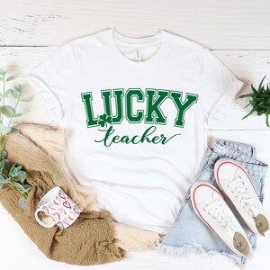 K&ouml;nnte beinhalten: Wei&szlig;es T-Shirt mit gr&uuml;nem Text "LUCKY teacher" und einem Kleeblatt-Motiv.