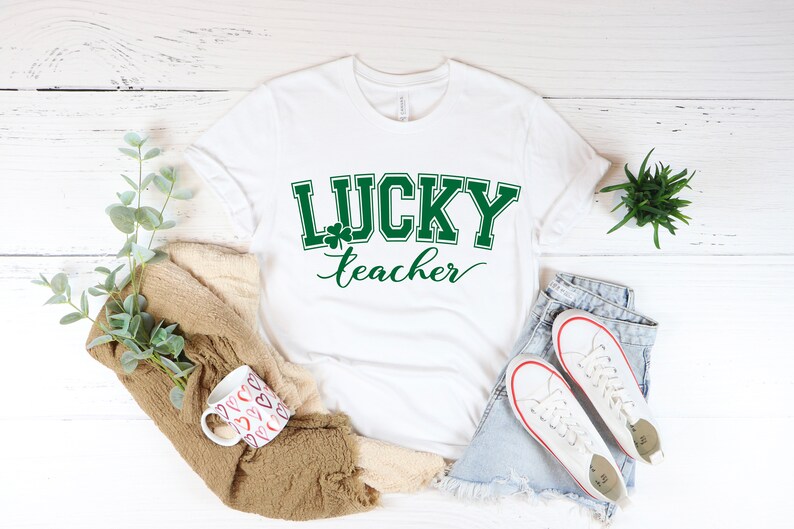 K&ouml;nnte beinhalten: Wei&szlig;es T-Shirt mit gr&uuml;nem Text "LUCKY teacher" und einem Kleeblatt-Motiv.