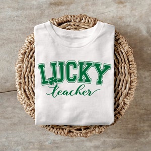 K&ouml;nnte beinhalten: Wei&szlig;es T-Shirt mit gr&uuml;nem Text, der "LUCKY teacher" lautet, mit einem Kleeblatt-Motiv.